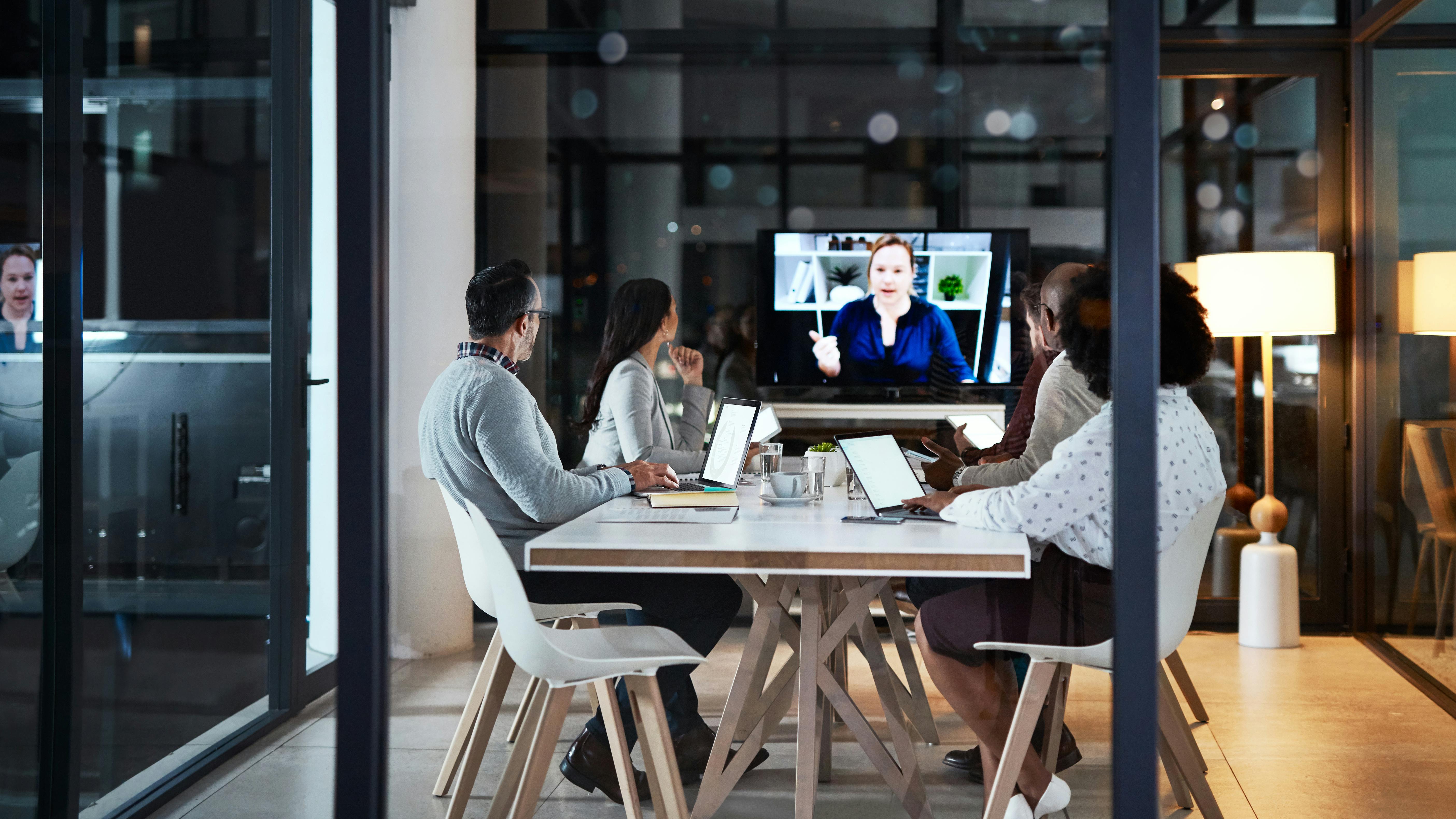 Modern workplace meeting room with people sitting around a conference table interacting with person on video screen.
