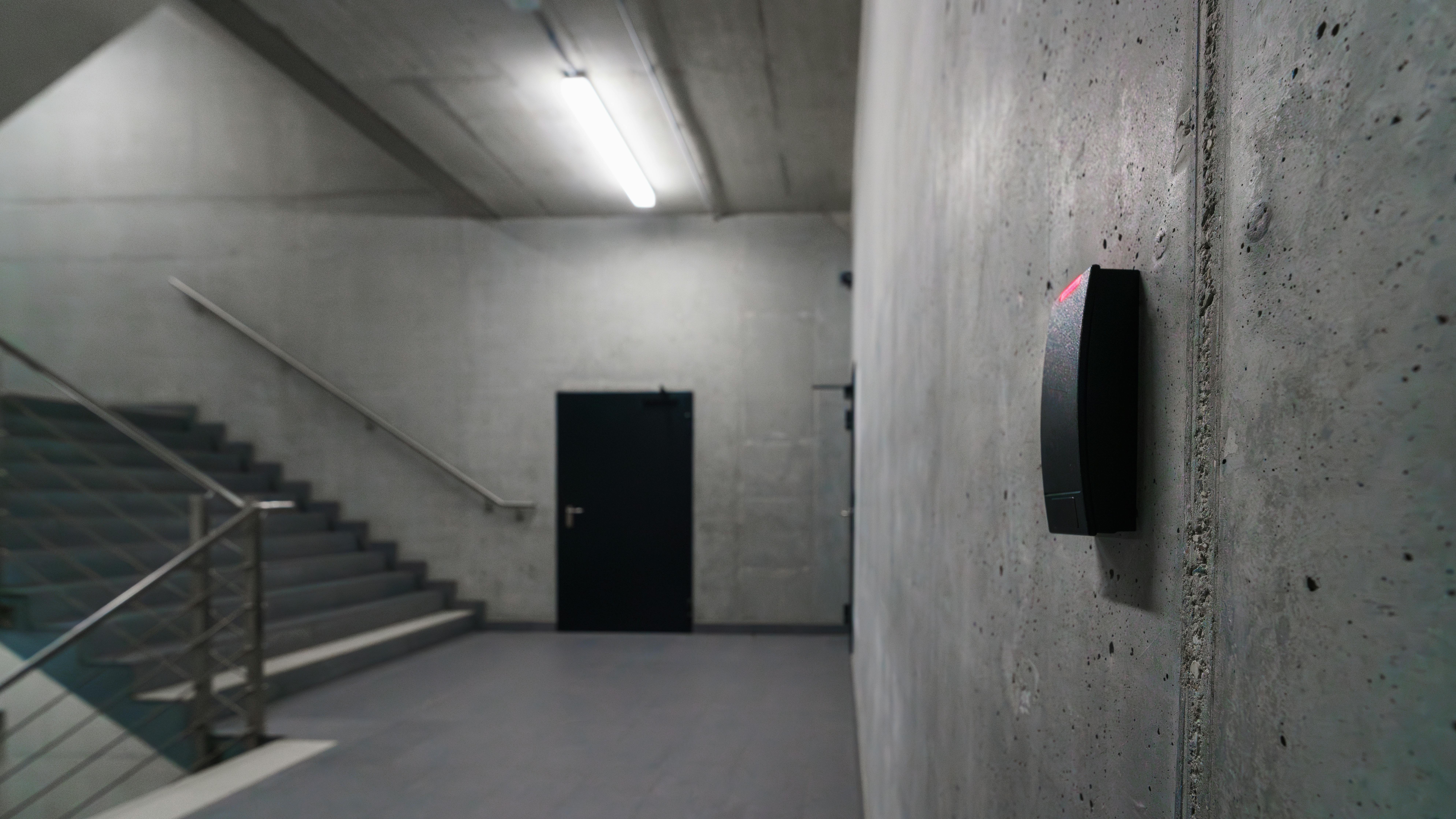 Concrete building stairwell with wall-mounted sensor in foreground and door in background.