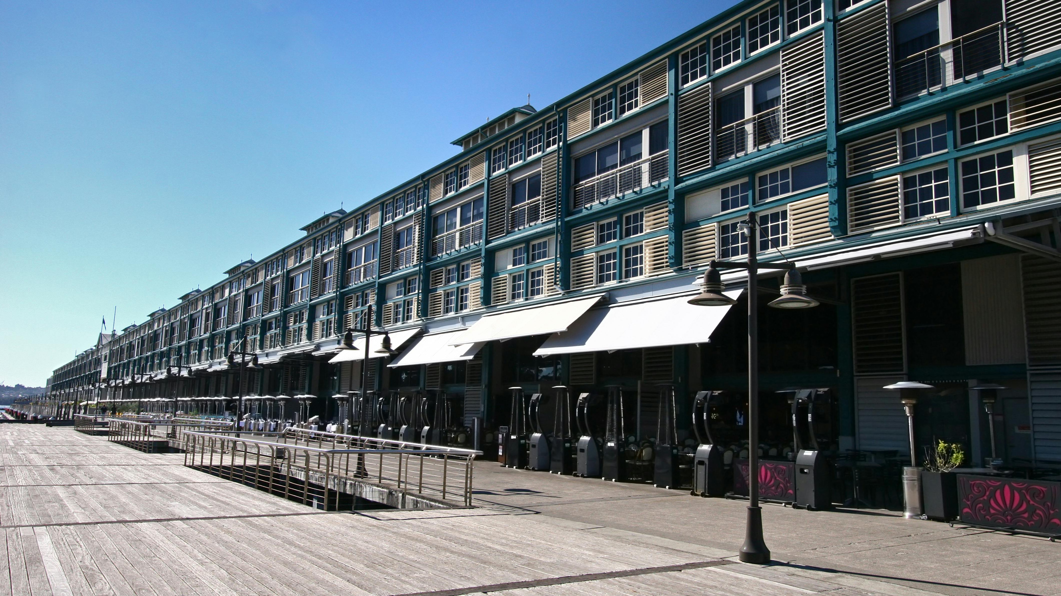 An adaptive reuse of a historic industrial wharf building at Finger Wharf, Woolloomooloo in Sydney, Australia. The heritage timber structure, featuring a green frame, white shutters, and louvers, has been transformed into the Ovolo Hotel, restaurants, and residential apartments, blending history with modern waterfront living.